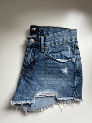 Blå jeansshorts  - Säljer ett par blå jeansshorts från 157 med rå, fransig kant och slitna detaljer. Shortsen har klassisk femficksdesign, knappgylf och asymmetrisk nederkant för en trendig look. Perfekta för varma dagar och enkel att matcha.