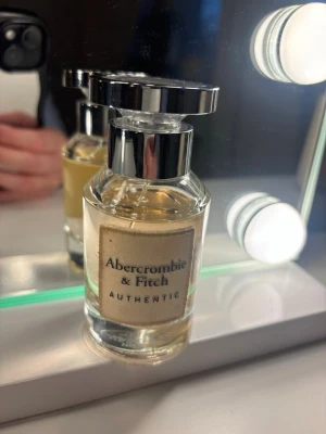 Abercrombie & Fitch Authentic parfym - En stilren parfym från Abercrombie & Fitch med transparent glasflaska och silverfärgat lock. Flaskan har en beige etikett med svart text och känns både modern och klassisk. Perfekt för dig som gillar fräscha och tidlösa dofter.
