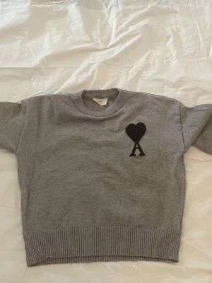 Grå stickad tröja från Ami Paris - Säljer en grå stickad tröja från Ami Paris med svart hjärta och bokstaven A broderat på bröstet. Tröjan har rund halsringning, ribbade muddar och nederkant. Perfekt för dig som gillar stilrena och trendiga plagg.