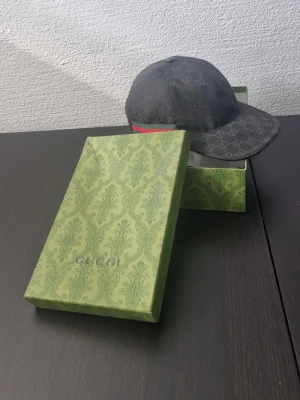 Svart Gucci keps canvas - Snygg svart keps från Gucci med klassiskt GG-monogram över hela kepsen och en grön-röd rand på sidan. Justerbar rem baktill i läder och metallspänne. Kepsen är tillverkad i bomull och levereras i originalförpackning.