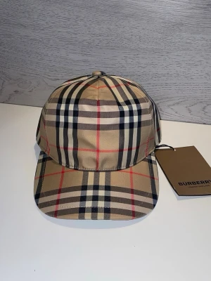 Beige keps med Burberry-rutor - Säljer en klassisk keps från Burberry i beige med det ikoniska rutmönstret i svart, vitt och rött. Kepsen har böjd skärm och justerbar rem bak. Tillverkad i ett slätt tyg som ger en stilren look. Perfekt för dig som gillar exklusiva accessoarer.