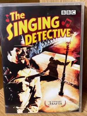 The Singing Detective DVD-box - Säljer DVD-boxen 'The Singing Detective', en brittisk kriminalthriller skapad av Dennis Potter. Serien blandar kriminalberättelse med musiknummer och självbiografiska inslag. Omslaget är färgstarkt med illustrationer och BAFTA-vinnare nämns. Perfekt för dig som gillar klassiska brittiska serier.