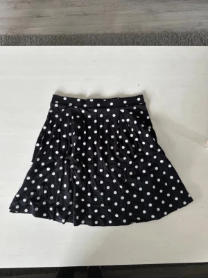 Svart prickig kortkjol - Säljer en svart kortkjol med vita prickar. Kjolen har en utställd form och bred linning i midjan. Perfekt för dig som gillar retrovibbar och vill ha en lekfull look.