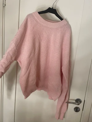 Ljusrosa oversized stickad tröja - Mysig ljusrosa stickad tröja med oversized passform och bred ribbad halsringning. Tröjan har långa ärmar och ribbade muddar vid ärmslut och nederkant. 