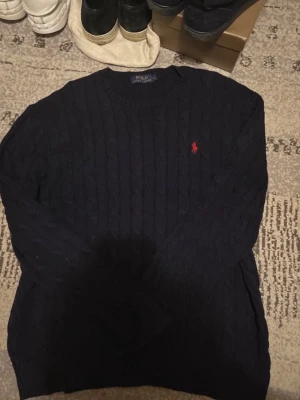 Mörkblå kabelstickad tröja Polo Ralph Lauren - Kabelstickad mörkblå tröja från Polo Ralph Lauren med rund halsringning och den klassiska röda logotypen broderad på bröstet. Tröjan har långa ärmar och ribbade muddar vid ärmslut och nederkant. Perfekt för en stilren och tidlös look.