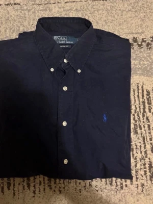 Mörkblå skjorta från Polo Ralph Lauren - Klassisk mörkblå skjorta från Polo Ralph Lauren med button-down krage och vita knappar. Skjortan har en broderad blå logga på bröstet och är tillverkad i bomull. Perfekt för en clean och stilren look. Pris kan diskuteras 