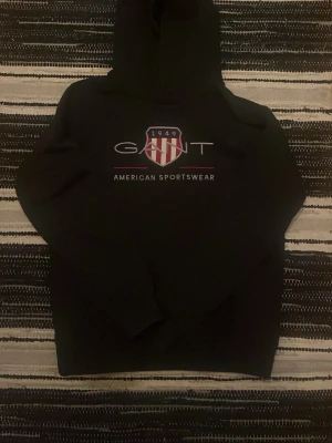 Svart hoodie från Gant med tryck - Säljer en svart hoodie från Gant med stort tryck på bröstet där det står 'GANT 1949 American Sportswear' och en sköld i rött och vitt. Klassisk huva och mjukt material, perfekt för en avslappnad och sportig stil.