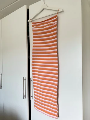 Randig långklänning i orange och vitt - Säljer en lång, ärmlös klänning med smala axelband och horisontella ränder i orange och vitt. Klänningen har en kroppsnära passform och är tillverkad i ett mjukt, stretchigt material som passar perfekt till sommaren.