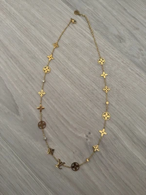 Guldigt halsband Louis Vuitton - Snyggt halsband i guldton från Louis Vuitton med ikoniska LV-symboler och blommor längs kedjan. Kedjan är tunn och elegant, dekorerad med små detaljer i guld och bruna nyanser. Perfekt accessoar för att lyfta din outfit.