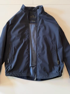 Paul & Shark Typhoon Jacket - Snygg mörkblå vindjacka från Paul & Shark med minimalistisk design. Jackan har hög krage, två framfickor med dragkedja och diskret logga. Jackans normalpris är 600 eur. Jackan är i prefekt kondition. Storleken är large me. Den passar också på storlek medium anvöndaren.
