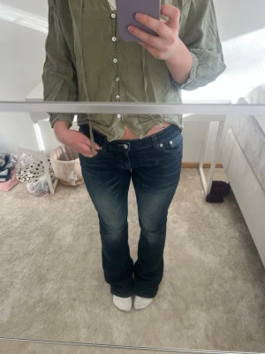 Mörkblå bootcut jeans med låg midja - Säljer ett par mörkblå bootcut jeans med låg midja och snygga slitningar framtill. Jeansen har klassiska fem fickor och silverfärgade nitar. Perfekta för en avslappnad och trendig look.