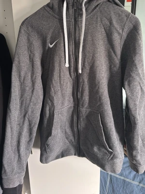Grå zip hoodie från Nike - Snygg grå hoodie från Nike med dragkedja framtill och vit logga på bröstet. Tröjan har huva med vita snören och två stora fickor framtill. Tillverkad i mjukt bomullsmaterial som är skönt att ha på sig. Perfekt för en avslappnad och sportig stil.