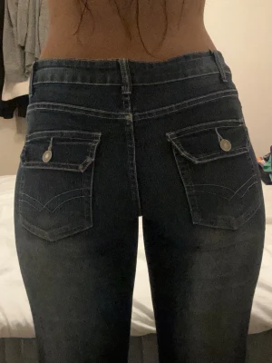 Mörkblå low waits bootcut jeans - Snygga mörkblå bootcut jeans med stora bakfickor med lock och knappdetaljer. Jeansen har klassisk femficksdesign och är tillverkade i stretchigt denimtyg för en bekväm passform. Perfekta för en trendig och avslappnad look.