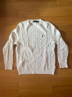 Vit kabelstickad tröja Polo Ralph Lauren - Kabelstickad vit tröja från Polo Ralph Lauren med klassisk rund halsringning och ribbade muddar. Tröjan har den ikoniska mörkblå logotypen broderad på bröstet och är tillverkad i mjukt material. Perfekt för en clean och stilren look.