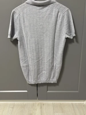 Ljusgrå ribbad polotopp John Henric  - Snygg ljusgrå polotopp från john Henric med ribbad struktur och korta ärmar. Toppen har klassisk krage och v-ringad öppning framtill. Perfekt till jeans eller kjol för en clean look.