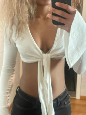 Vit croppad omlottopp med knyt - Säljer en vit croppad omlottopp med långa ärmar och knyt framtill. Toppen har djup v-ringning och är perfekt för dig som vill ha en trendig och stilren look. Passar till både jeans och kjol för en cool vibe. Den har ett till hål/remma där man kan knyta ifall det behövs 
