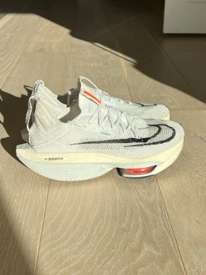 Nike ZoomX Vit Flyknit springskor - Nike ZoomX sneakers i vitt Flyknit-material med svart Swoosh på sidan och orange detalj på plösen. Skorna har en chunky sula med synlig dämpning och modern design, perfekta för dig som gillar sportig och futuristisk stil.