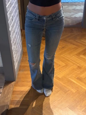 Blå bootcut jeans med slitningar - Säljer ett par ljusblå bootcut jeans med slitningar på benen och broderade fickor bak. Jeansen har låg midja och klassisk femficksmodell. Materialet är denim i bomull och de har en avslappnad, trendig vibe. Passar som de vore xs