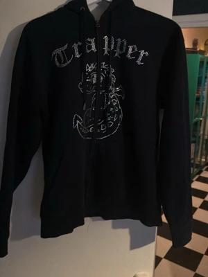 Svart Trapper hoodie med drakmotiv - Säljer en svart hoodie från Trapper med dragkedja och huva. På framsidan finns ett stort tryck med en tecknad drake och texten 'Trapper' i glittriga bokstäver. Hoodien har långa ärmar och snörning i huvan. Perfekt för dig som gillar streetwear och coola prints.