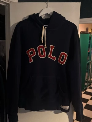 Marinblå hoodie från Polo Ralph Lauren - Snygg marinblå hoodie från Polo Ralph Lauren med stor röd och vit POLO-text på bröstet. Klassisk huva med vita dragsnören och känguruficka framtill. Tillverkad i mjuk bomullsblandning, perfekt för en avslappnad och sportig stil.