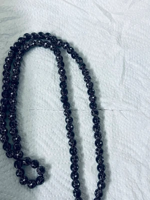 Svart pärlhalsband med silverdetaljer misabaha  - Snyggt svart halsband med runda pärlor och silverfärgade detaljer. Halsbandet har en dekorativ tofs i änden med flätade trådar och extra pärlor. Perfekt accessoar för att lyfta din outfit med en unik touch.