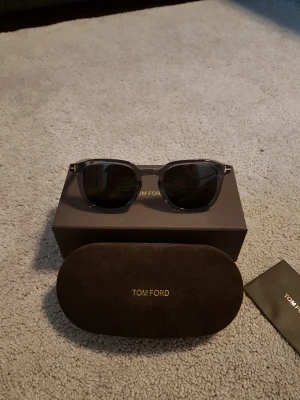 Transparenta solglasögon från Tom Ford - Snygga transparenta solglasögon från Tom Ford med klassisk fyrkantig form och mörka linser. Bågarna är tillverkade i syntetmaterial och har diskreta metallinslag vid sidorna. Kommer med originalfodral och box.