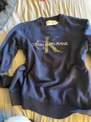 Mörkblå sweatshirt Calvin Klein Jeans - Mörkblå sweatshirt från Calvin Klein Jeans med stort CK-logotryck i silver på bröstet. Tröjan har rund halsringning, ribbade muddar vid ärmslut och nederkant samt en avslappnad passform. Tillverkad i mjukt bomullsmaterial för skön känsla.