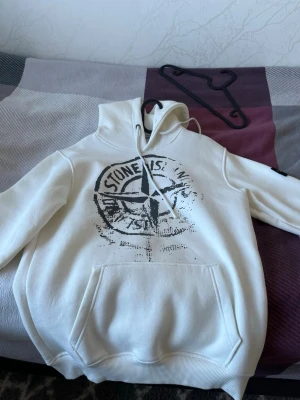 Vit hoodie från Stone Island - Säljer en vit hoodie från Stone Island med stor logga i svart på bröstet. Hoodien har huva med dragsko och en stor magficka framtill. Tillverkad i mjukt material som känns skönt mot huden. Perfekt för dig som gillar streetwear och vill ha en clean look. Helt ny och riktig, pris kan diskuteras 
