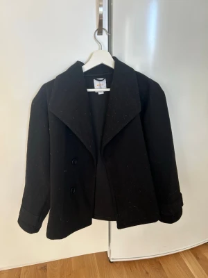 Svart kappa från Gina Tricot - Stilren svart kappa från Gina Tricot med dubbelknäppning och bred krage. Klassisk rak modell med uppvikta ärmslut, perfekt för en trendig look. Tillverkad i ett mjukt tyg som ger en snygg siluett. Passar perfekt till höstens outfits.