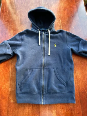 Marinblå hoodie från Polo Ralph Lauren - Snygg marinblå hoodie från Polo Ralph Lauren med dragkedja, två fickor framtill och klassisk gul logga på bröstet. Mjuk bomullskänsla, huva med vita snören och ribbade muddar. Perfekt för en chill och stilren look. Sitter som en S