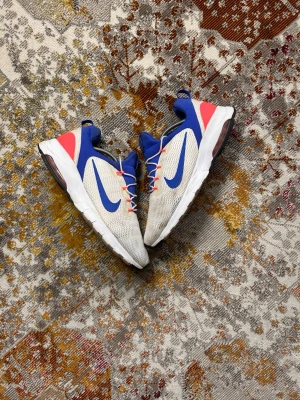 Nike Air Max vita/blå sneakers - Nike Air Max sneakers i vitt meshmaterial med blå detaljer och stor blå Swoosh på sidan. Skon har inslag av rött vid hälen och vita snören med röda accenter. Synlig Air Max-dämpning i sulan och svart yttersula. Perfekt för dig som gillar sportig och modern stil.