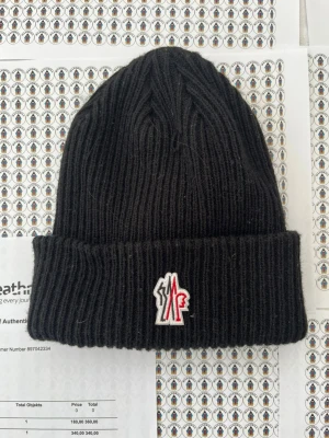 Svart ribbstickad mössa Moncler - Svart ribbstickad mössa från Moncler med uppvikt kant och ikonisk logotyp framtill i vitt, rött och svart. Klassisk passform och mjukt material som håller dig varm. Perfekt accessoar för kyliga dagar.