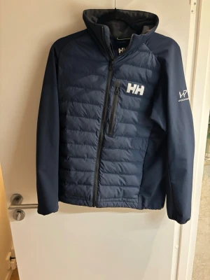 Mörkblå pufferjacka från Helly Hansen - Snygg mörkblå pufferjacka från Helly Hansen med hög krage, dragkedja framtill och bröstficka med dragkedja. Jackan har HH-logga på bröstet och ryggen samt HP-logga på ärmen. Tillverkad i slitstarkt material med vadderade paneler för extra värme. Jackan är väldigt fet och svår att få tag på ny!
