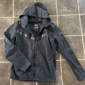 Svart Nike Tech Fleece - Snygg svart tech fleece från Nike med dragkedja framtill och huva. Den har diskreta svarta detaljer över bröstet och en praktisk ficka med dragkedja på ena ärmen. Tillverkad i ett mjukt och bekvämt material, perfekt för en avslappnad stil. Superbra kvalité, nästan helt oanvänd. Postar inom 1-2 dagar. Pris ej hugget i sten så kom med bud.