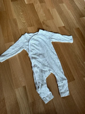 Vit babypyjamas från Primark - Mysig vit babypyjamas från Primark med diskret mönster och omlottknäppning framtill. Tillverkad i mjukt tyg, perfekt för sovstunden. Långa ärmar och ben med tryckknappar längs benet för enkel påklädning.