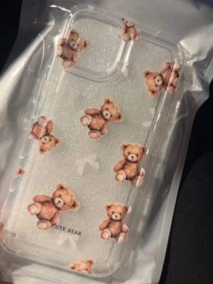 Genomskinligt mobilskal med nallemotiv - Säljer ett genomskinligt mobilskal dekorerat med söta bruna nallar och små vita rosetter. Skalets design är lekfull och trendig, perfekt för dig som vill ha ett gulligt och personligt skydd till mobilen. Texten 'CUTE BEAR' finns längst ner på skalet.
