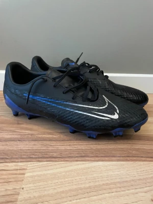 Nike Fotbollsskor Phantom - Säljer ett par svarta Nike fotbollsskor med blå och silverfärgade detaljer. Skorna har klassisk snörning, låg profil och dobbar för spel på gräs. Tillverkade i syntetmaterial för bra grepp och kontroll på planen. Perfekta för dig som vill ha stilrena och funktionella skor. Storlek 44 Phantom GX