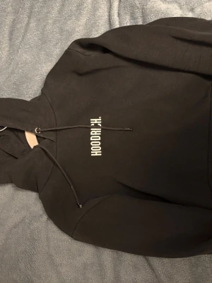 Svart hoodie från Hoodrich - Säljer en svart hoodie från Hoodrich med vit logga tryckt vertikalt på bröstet. Hoodien har klassisk huva med dragsko och är tillverkad i mjukt bomullsmaterial. Perfekt för dig som gillar streetwear och vill ha en enkel men snygg look.