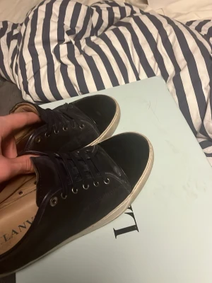 Svarta Lanvin sneakers i läder - Snygga svarta sneakers från Lanvin i läder med vita sulor och svarta skosnören. Klassisk låg modell med metallöljetter och diskret branding på insidan. Perfekta för en clean och stilren look.