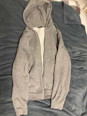 Grå zip hoodie med huva - En klassisk grå zip hoodie från H&M med huva och dragsko. Hoodien har två stora fickor framtill och ribbade muddar vid ärmslut och nederkant. Insidan är mjuk och borstad för extra komfort. Perfekt för en avslappnad och chill stil.