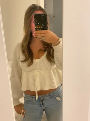 Vit cropped stickad tröja med knyt - Supersöt vit stickad tröja med croppad passform och knytband i midjan. Tröjan har långa ärmar, v-ringad hals och volangdetalj nedtill. Perfekt att styla med jeans för en trendig look.