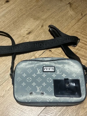 Grå axelväska från Louis Vuitton - Snygg grå axelväska från Louis Vuitton med klassiskt monogrammönster och svart justerbar axelrem med logotyp. Väskan har en svart ficka framtill med spänne och detaljer i silver. Perfekt för dig som vill ha en trendig och praktisk accessoar.