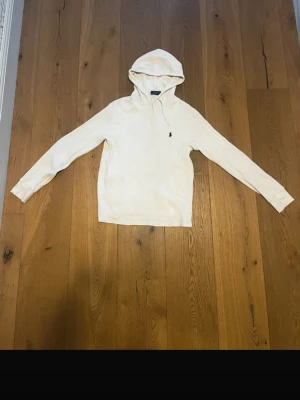 Vit hoodie från Polo Ralph Lauren - Säljer en stilren vit hoodie från Polo Ralph Lauren med klassisk huva och dragsnören. Tröjan har en broderad logga på bröstet och en stor magficka framtill. Tillverkad i mjukt material som känns skönt mot huden. Perfekt för en avslappnad look.