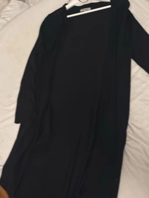 Svart lång kofta från Ullared  - Stilren svart lång kappa från Ullared  med öppen front och raka ärmar. Perfekt för dig som gillar minimalistisk och enkel design. Kappan har en rak passform och är tillverkad i ett mjukt tyg som känns skönt att bära.