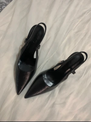 Svarta spetsiga klackar med rem - Säljer ett par svarta pumps med spetsig tå och smal klack. Skorna har en elegant rem runt hälen och är tillverkade i ett glansigt material som liknar skinn. Dem är helt nya och oanvända som ni ser. Perfekta för dig som gillar en klassisk och stilren look.