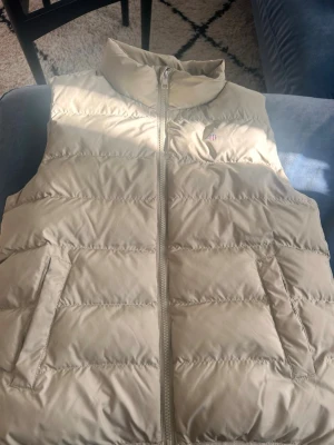 Beige dunväst från Gant 146/152 - Snygg beige dunväst från Gant med hög krage och dragkedja framtill. Västen har två fickor med lock och. Köpt i NK på gant affären, nypris runt 1400 säljer pga för liten och växt ur den. Perfekt till lite kyliga sommmar kvällar 
