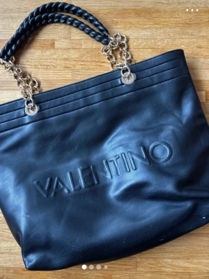 Svart shoppingväska från Valentino - Snygg svart shoppingväska från Valentino i mjukt konstläder med präglad logga framtill. Väskan har tvinnade handtag i svart och guldfärgade kedjor som detaljer. Perfekt storlek för att få plats med allt du behöver.