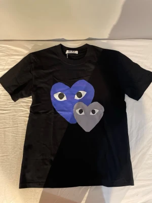 Svart Comme des Garçons t-shirt - Svart t-shirt från Comme des Garçons med två stora hjärtan på bröstet, ett blått och ett grått, båda med ögonmotiv. Klassisk passform och rund halsringning. Perfekt för dig som gillar streetwear och grafiska prints.