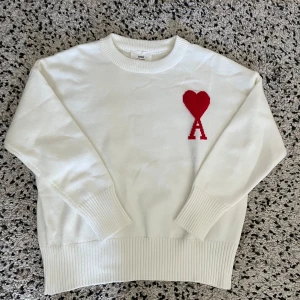Vit stickad tröja från Ami Paris - Vit stickad tröja från Ami Paris med röd hjärtlogga och bokstaven A på bröstet. Tröjan har rund hals, ribbade muddar och är långärmad. Perfekt för dig som gillar stilrena och trendiga plagg med en twist.