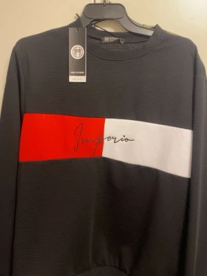 Svart tröja med röd och vit panel - Cool svart tröja från IAM Studio med bred röd och vit panel över bröstet och texten 'Imperio' broderad i svart. Tröjan har rund halsringning och ribbade muddar. Perfekt för en trendig och avslappnad stil.